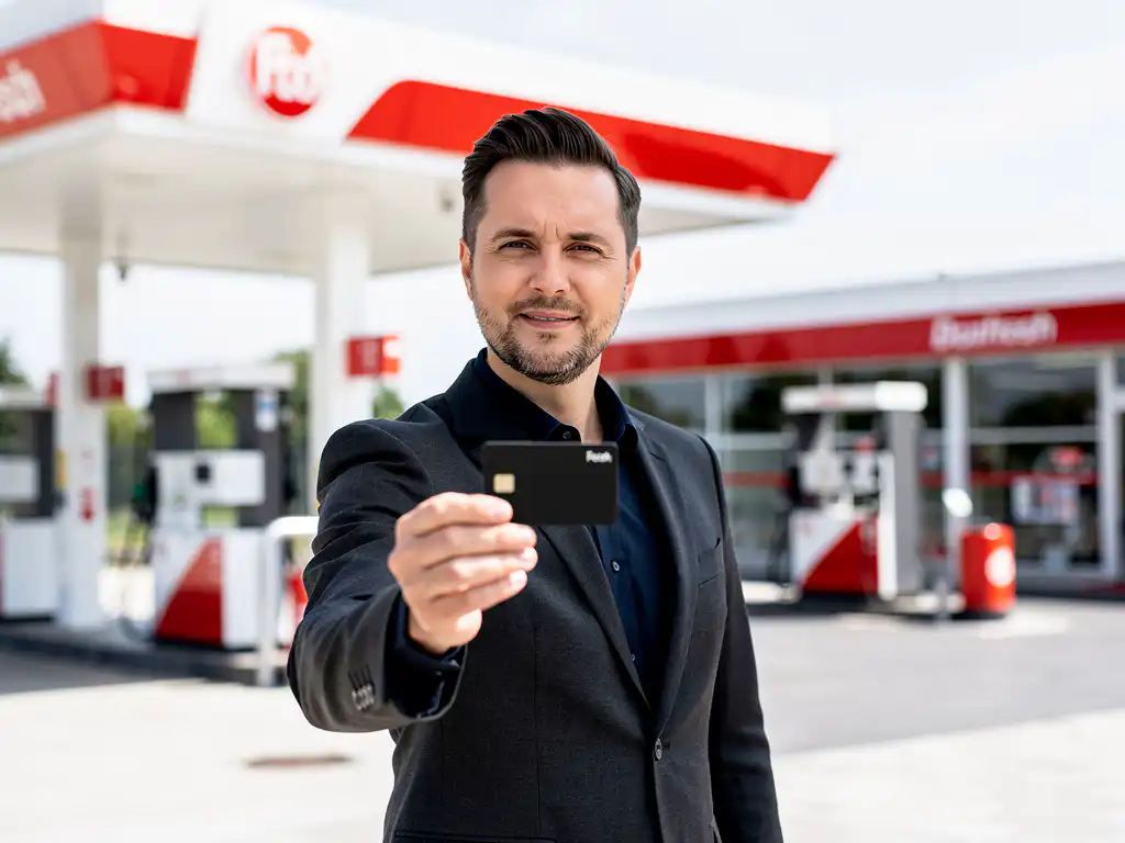 Ondernemer in donker blazer houdt zwarte brandstofpas vast bij modern Nederlands tankstation met wit-rood luifel.
