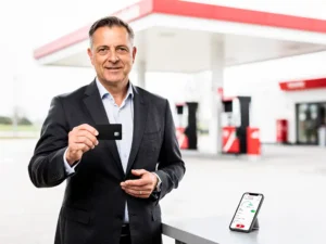 Zakenman in donker blazer houdt tankpas vast bij modern Nederlands tankstation met smartphone waarop kilometerregistratie-app zichtbaar is.