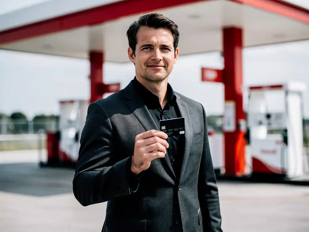 Zakenman in donker blazer houdt tankpas vast bij modern Nederlands tankstation met wit-rode huisstijl.