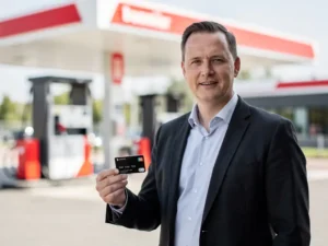 Vlootbeheerder in donker blazer houdt brandstofpas vast bij modern Nederlands tankstation met wit-rood luifel.