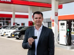 Vlootbeheerder met zwarte brandstofpas bij Nederlands tankstation met laadpaal, brandstofpomp en bedrijfsvoertuigen op de achtergrond.