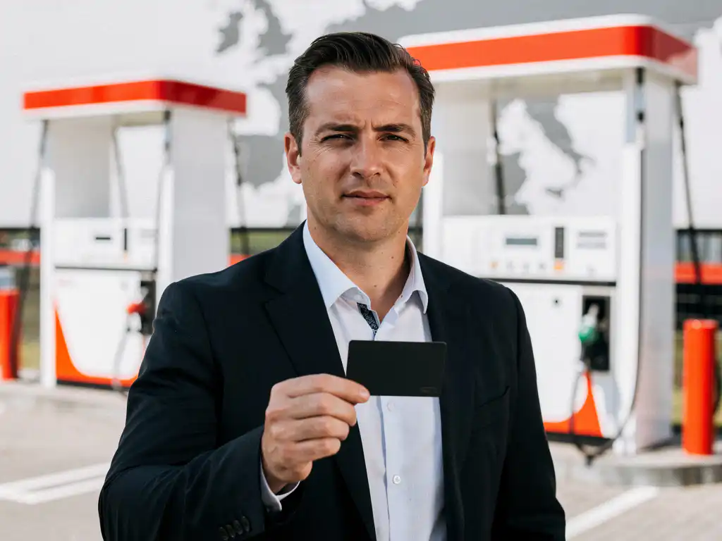 Wagenparkeigenaar in donker blazer met brandstofpas bij modern Nederlands tankstation met witte en rode pompoverkappingen.
