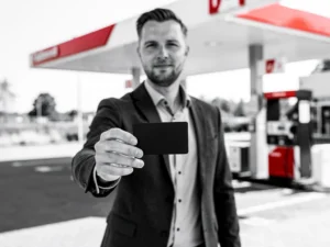 Zelfstandige ondernemer in donker blazer houdt brandstofpas vast bij modern Nederlands tankstation met rood-wit luifel.